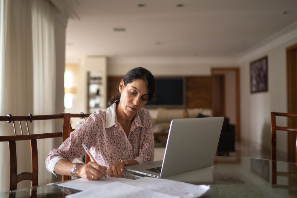 A imagem possui uma mulher sentada em uma mesa, com um notebook e alguns papeis, dando ao entender que esteja trabalhando em home office.