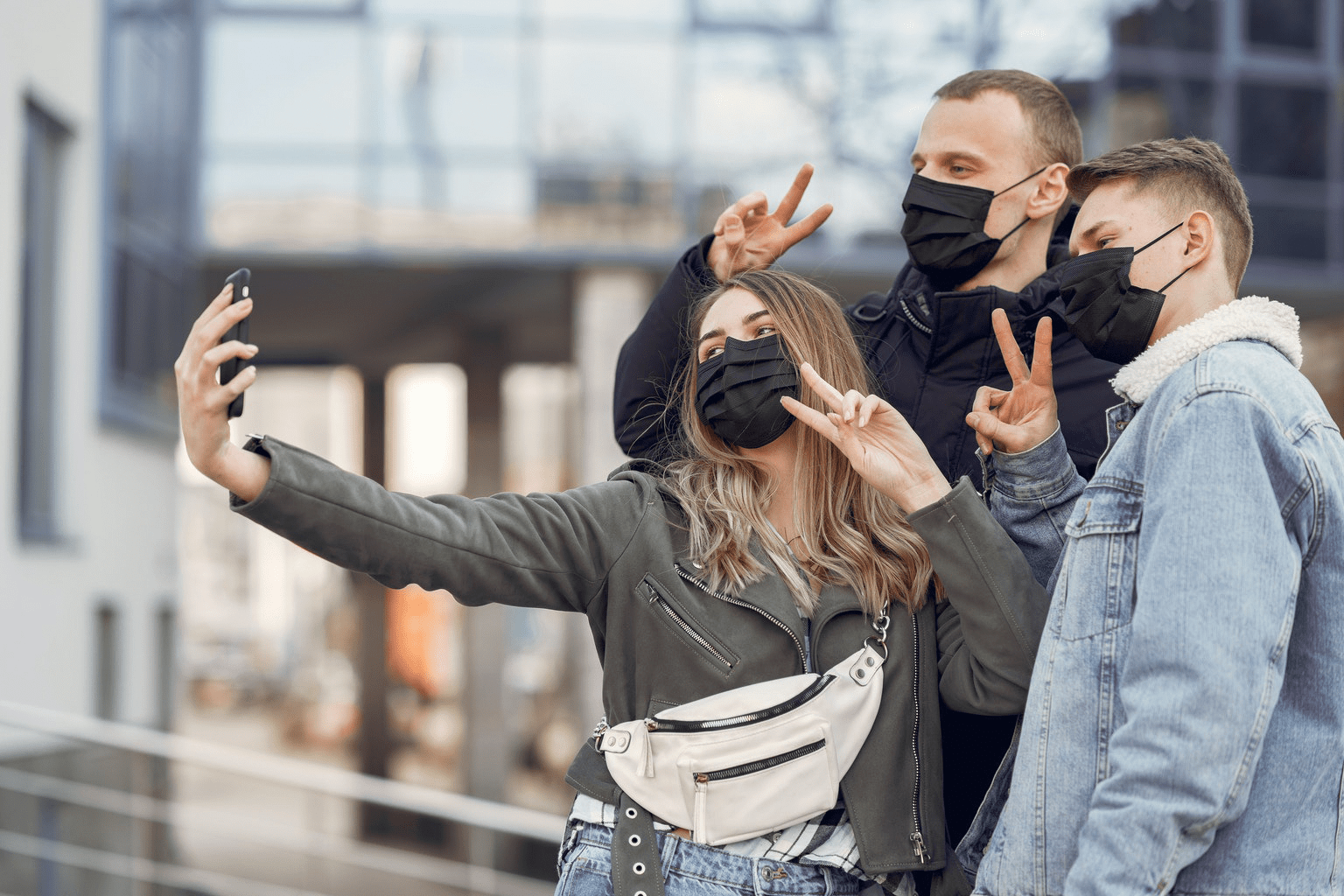 três jovens, sendo uma mulher e dois homens, usando mascarás de proteção contra o coronavírus, fazendo sinal de paz e amor com as mãos e olhando para a câmera da mulher para tirar uma foto estilo selfie