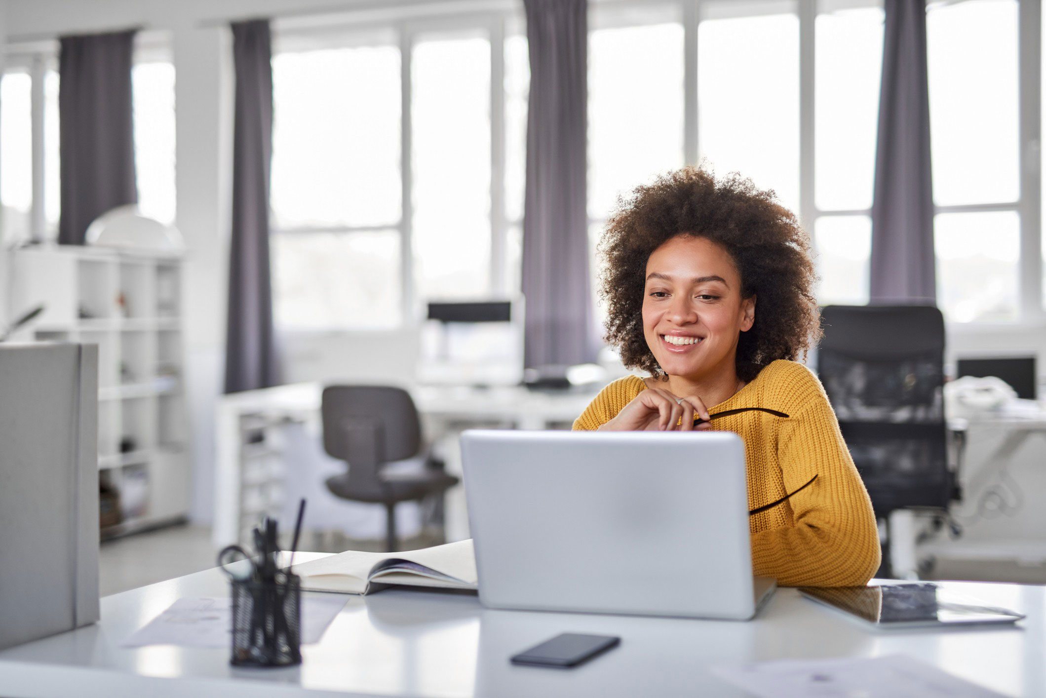 Mulher, parda, com blusa amarela, usando um notebook em um escritório.