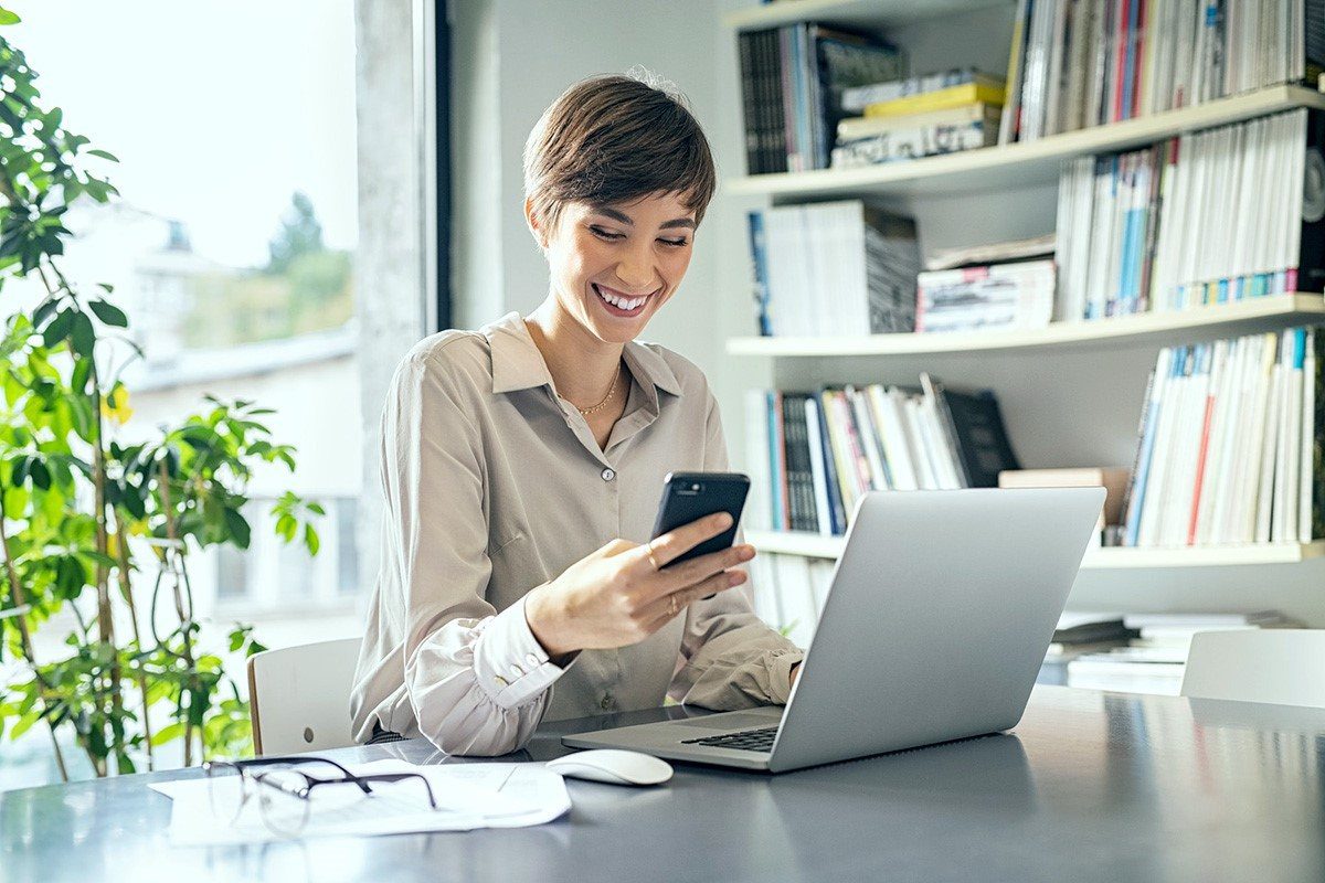 Mulher de cabelo curto feliz no escritório olhando para tela do smartphone e com notebook na sua frente